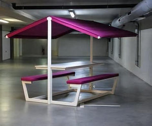A modern outdoor dining set consisting of a wooden table and two matching benches with magenta-purple cushions, under a matching purple canopy, set in a large concrete indoor patio area.