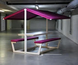 A modern outdoor dining set consisting of a wooden table and two matching benches with magenta-purple cushions, under a matching purple canopy, set in a large concrete indoor patio area.