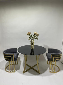 A modern 3-piece dining set featuring a round black-top table with a gold metal base, flanked by two gray velvet armchairs with gold geometric metal frames, styled with a gold vase of flowers in the center.