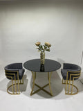 A modern 3-piece dining set featuring a round black-top table with a gold metal base, flanked by two gray velvet armchairs with gold geometric metal frames, styled with a gold vase of flowers in the center.
